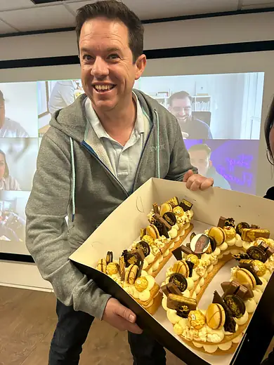 A colleague holding up a tray of pastries in front of a video call full of faces.