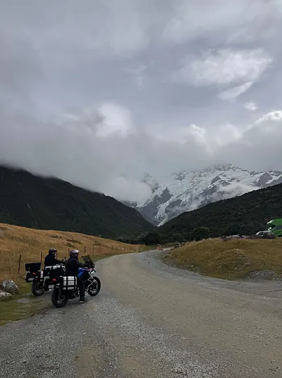 Two motorcycles on a gravel road below a snow-covered mountain.