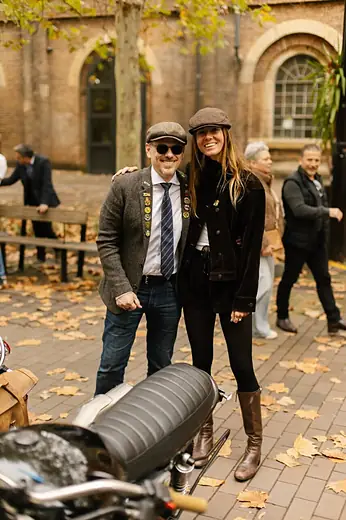 Two people in flat caps beside a motorcycle on an autumn square.